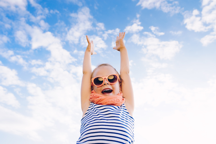 Happy beautiful child girl in sunglasses with raised arms against the sky